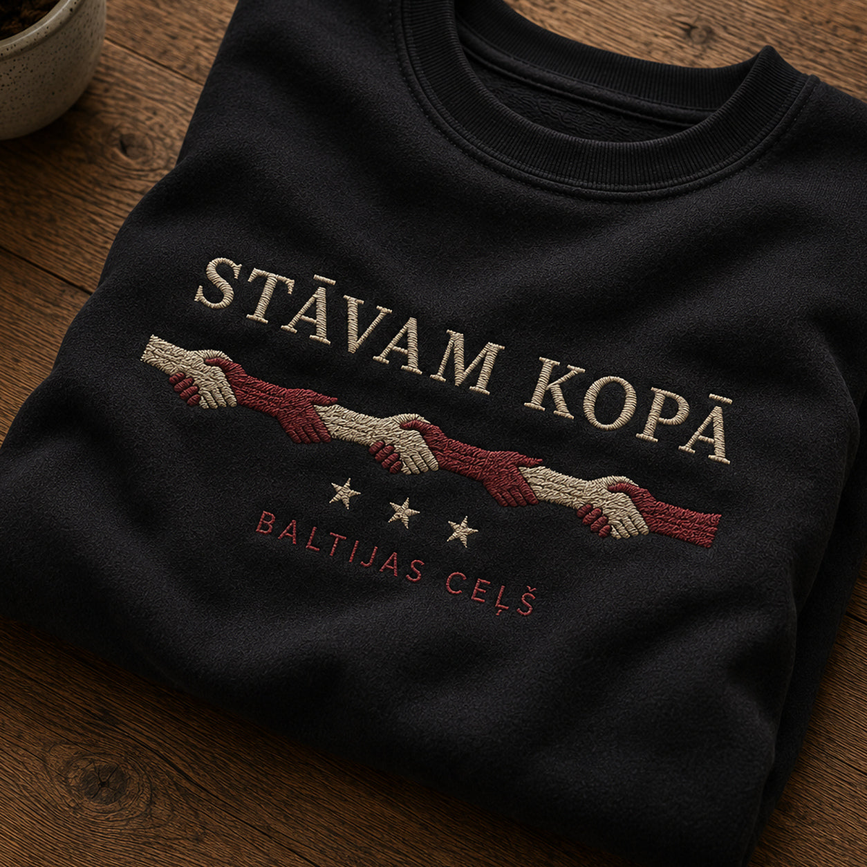 Tuvāks “Stāvam kopā” kompozīcijas skats uz melnu salocītu džemperi ar tekstu “Stāvam kopā” un apakštekstu “Baltijas ceļš”