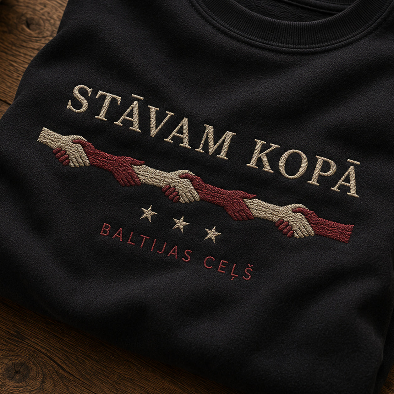 “Stāvam kopā” izšuvuma detaļu tuvplāns uz melna džempera auduma, redzams motīvs: vienotības aicinājums Stāvam kopā un vēsturiskais nosaukums Baltijas ceļš