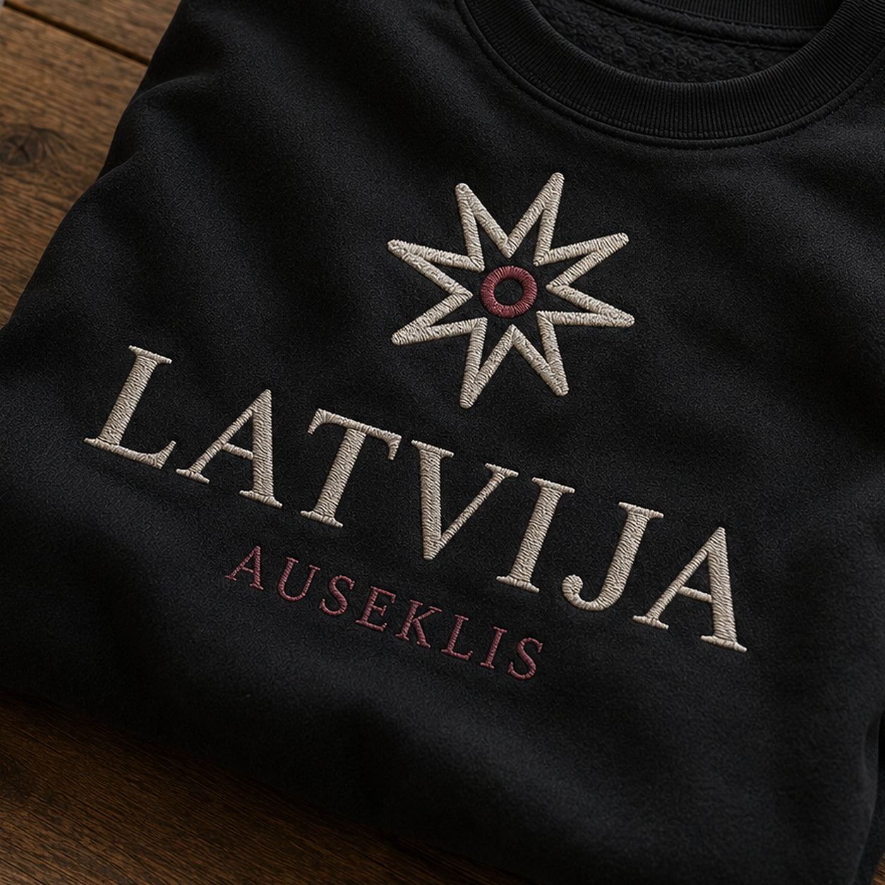 “Auseklis Latvija” izšuvuma detaļu tuvplāns uz melna džempera auduma, redzams motīvs: Ausekļa astoņstaru ritms un skaidrais vārds Latvija